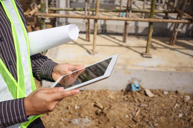 Construction manager and engineer worker in protective helmet hold blueprints paper on hand using tablet computer at building site