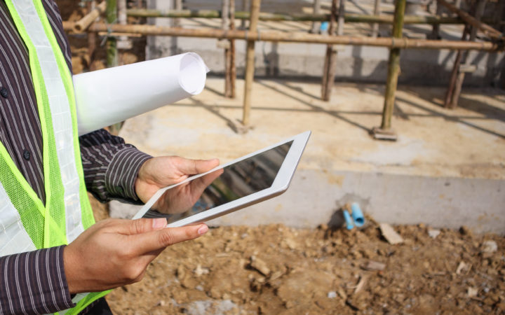 Construction manager and engineer worker in protective helmet hold blueprints paper on hand using tablet computer at building site